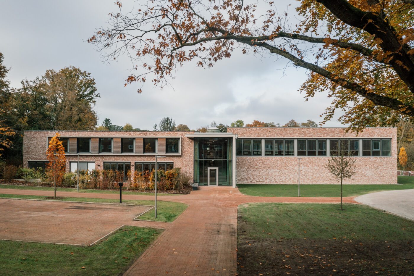 Modernes Backsteingebäude mit Glasfassade in herbstlicher Landschaft, umgeben von Bäumen und Parkwegen