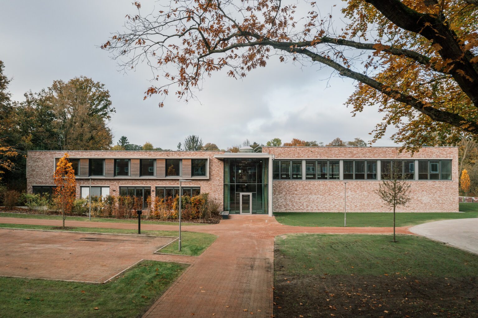 Modernes Backsteingeb&auml;ude mit Glasfassade in herbstlicher Landschaft, umgeben von B&auml;umen und Parkwegen