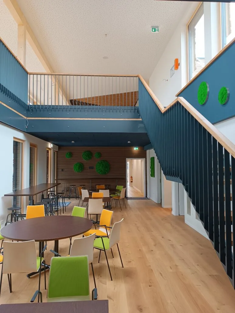 Ein moderner, heller Innenraum mit Holzboden und zweistöckigem Grundriss. Der Raum verfügt über eine Treppe mit dunklem Geländer, runde Tische mit bunten Stühlen und grüne Pflanzendekorationen an den Wänden. Natürliches Licht strömt durch große Fenster.