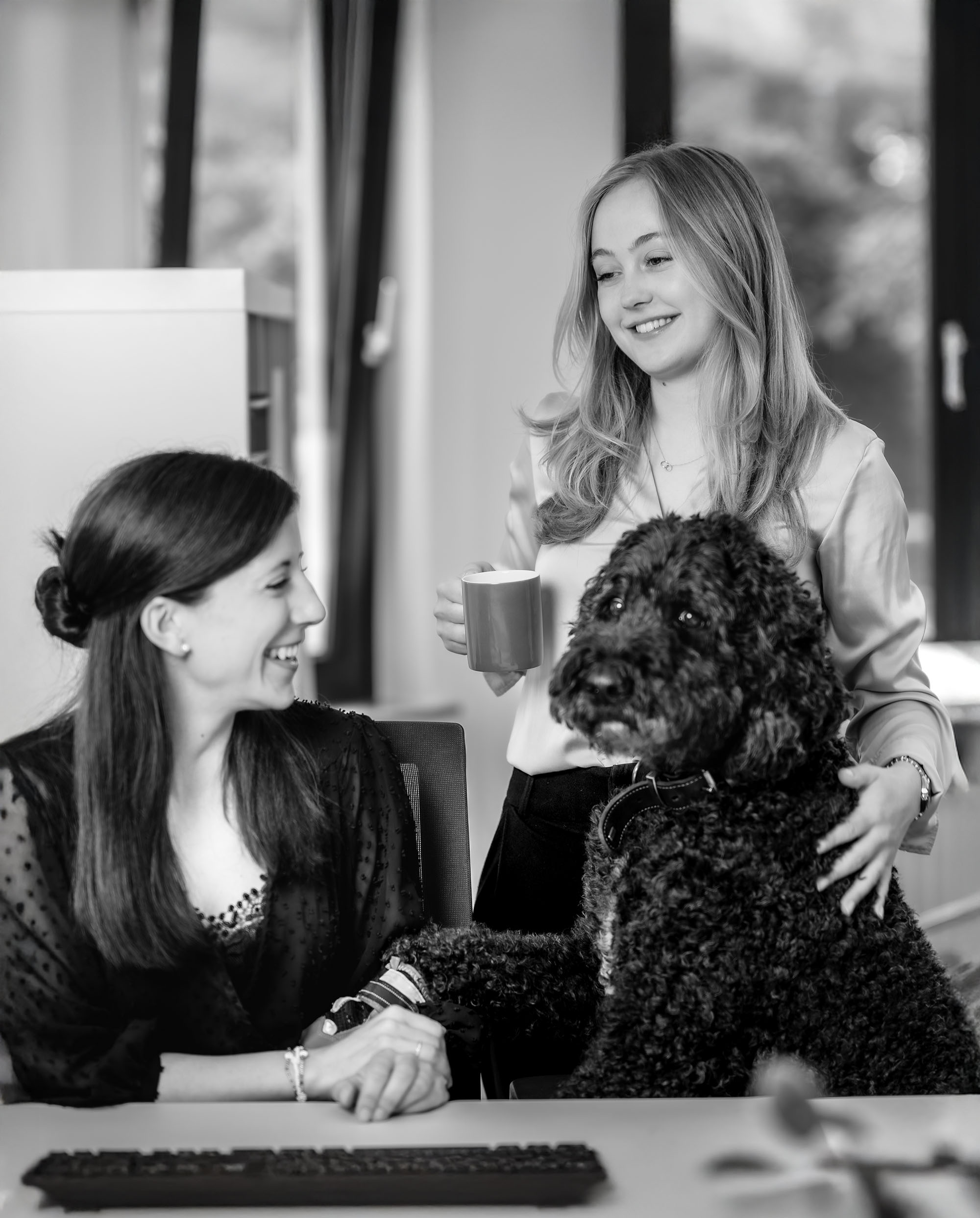 Zwei Frauen im Büro mit einem Hund, eine hält eine Tasse, die andere lächelt, während der Hund sie aufheitert