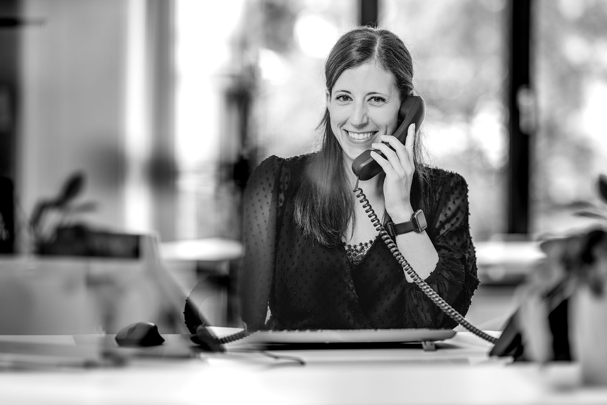 Frau im Büro lächelt am Telefon, schwarz-weiß Bild, arbeitet am Schreibtisch mit Laptop im Hintergrund