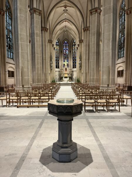 Innenansicht einer gotischen Kirche mit Taufbecken im Vordergrund und Altar sowie bunten Glasfenstern im Hintergrund