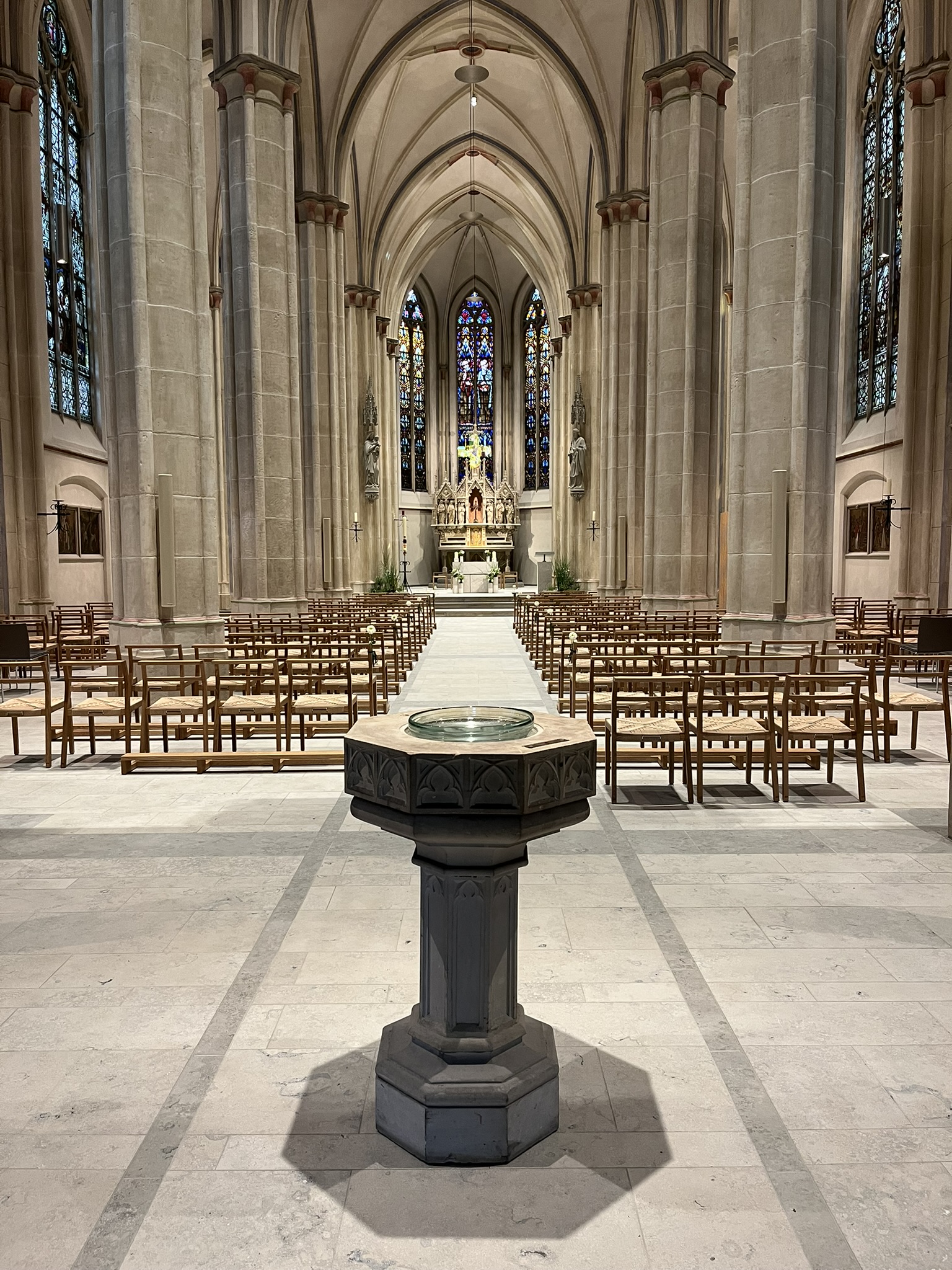 Innenansicht einer gotischen Kirche mit Taufbecken im Vordergrund und Altar sowie bunten Glasfenstern im Hintergrund