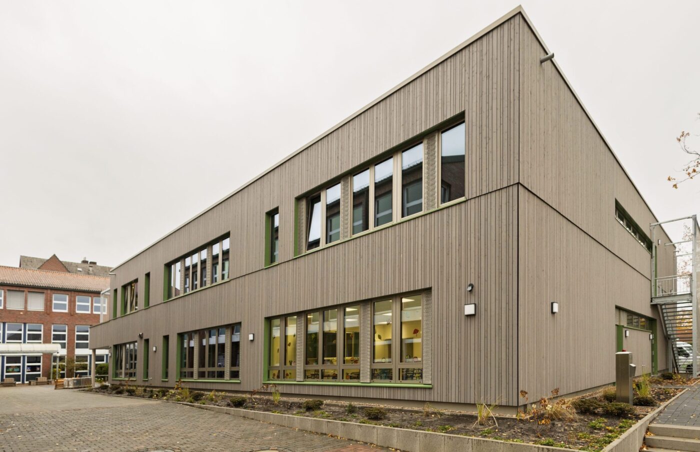 Moderne Schulgebäude mit großen Fenstern und Holzelementen, Ansicht des gepflasterten Pausenhofs und Eingangstreppen